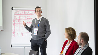 Ein Mann im grauen Pullover steht neben einem ausgefüllten Flipchart mit der roten Überschrift „EPR with Eco-Modulation“ und präsentiert den im Vordergrund sitzenden Zuhörenden gestikulierend die Ergebnisse.