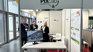Drei Teammitglieder des PtX Lab Lausitz an ihrem Messestand. Ein Monitor im Vordergrund zeigt Grafiken, an der Wand hängen Infografiken und das Logo des Labs ist groß auf der Standblende zu sehen.