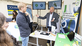 Ein Referent des PtX Lab Lausitz erklärt einem Besucher am Info-Stand die Wirkungsweise eine Elektrolyseurs.