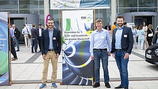 Dre Mitarbeiter der PtX Lab Lausitz stehen vor einem kleinen Plakat des Labs auf dem Gelände des Zentralcampus der Brandenburgischen Technischen Universität (BTU) Cottbus-Senftenberg. 