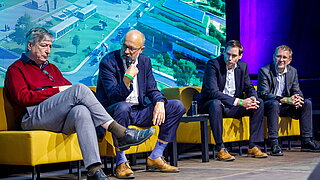 Vier Experten sitzen während einer Podiumsdiskussion bei den deCarbonDays auf gelben Sesseln. Ein Sprecher in der Mitte moderiert mit einem Mikrofon vor einer großen Videowand, die eine Industrieanlage zeigt.