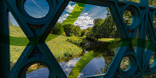 Blick durch die Freiflächen einer Maßwerkballustrade auf eine hügelige Wiesenlandschaft mit einem Bachlauf im Vordergrund und Bäumen im Hintergrund und durch grafische Nachbearbeitung darübergelegt die grünen gebogenen Linien aus dem Logo von PtXLab Lausitz