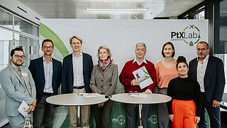 Ein Gruppenfoto von acht Personen vor einem großen weißen Banner mit dem PtX Lab Lausitz Logo in Berlin. In der Mitte hält ein Mann in einem roten Pullover ein gedrucktes Exemplar einer Studie hoch. Vor der Gruppe stehen zwei runde Bistrotische.