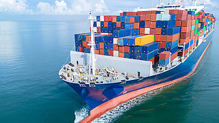 Aerial front view of a large cargo ship fully loaded with colorful shipping containers navigating through calm ocean water.