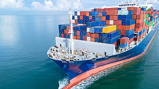 Luftaufnahme eines großen Frachtschiffs, das vollständig mit bunten Schiffscontainern beladen ist und durch ruhiges Meerwasser fährt.