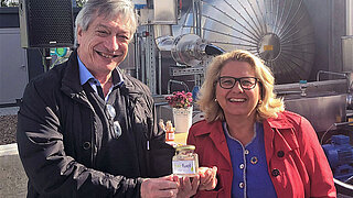 Harry Lehmann und Svenja Schulze stehen in einer Produktionsanlage unter freiem Himmel und halten ein kleines Gefäß mit der Aufschrift "fairfuel"