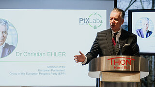 In the foreground, a man is standing behind a lectern, speaking. Behind him, a presentation is shown on a screen.