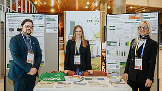 Drei Teammitglieder des PtX Lab Lausitz stehen lächelnd an ihrem Messestand bei der Nova-Konferenz 2025 in Köln. Hinter ihnen hängen wissenschaftliche Poster zu chemischer Produktion und E-Kerosin. Auf einem weiß gedeckten Tisch im Vordergrund liegt Informationsmaterial bereit.