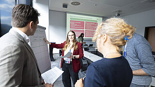 Eine Frau in einem roten Blazer zeigt bei der Renewable Materials Conference auf eine Tabelle an einem Flipchart und diskutiert mit umstehenden Teilnehmenden, während im Hintergrund eine Folie zu „Market-Pull Instruments“ des PtX Lab Lausitz projiziert wird.