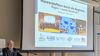 Ein Redner im Anzug spricht an einem Mikrofon vor einer großen Leinwand. Auf der Projektion ist der Titel „Wasserstofftour durch die Regionen: Region: Lausitz-Spreewald“ sowie Logos verschiedener brandenburgischer Ministerien und Organisationen zu sehen.