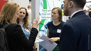 Eine Gruppe von Fachleuten im Gespräch am Stand des PtX Lab Lausitz auf der ILA Berlin. Eine Frau gestikuliert lebhaft, während sie Dokumente hält. Christin Heldt vom PtX Lab und weitere Teilnehmende hören aufmerksam zu. Im Hintergrund ist ein Roll-up-Banner des „Think and Do Tanks“ zu sehen.