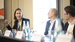 A woman is sitting at a table in a meeting room, speaking into a table microphone. Two men are listening.
