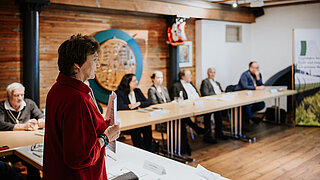 Eine Frau in einer roten Jacke steht im Vordergrund und hält Unterlagen, während sie zu einer Gruppe spricht, die an Tischen in einem rustikalen Raum sitzt. Im Hintergrund ist ein Banner des PtX Lab Lausitz zu sehen.