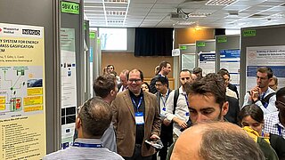 Eine belebte Poster-Session auf einer wissenschaftlichen Konferenz. In der Mitte steht ein lächelnder Experte des PtX Lab Lausitz in einem braunen Sakko. Fachbesucher betrachten technische Poster, unter anderem zu Biomassevergasung und Energiesystemen.