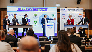 Podiumsdiskussion auf der aireg Biennial SAF Conference 2025 in Berlin. Fünf Experten stehen auf einer Bühne vor einer großen Leinwand mit dem Slogan „LET’S FLY SAF. NOW!“