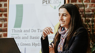 Porträtaufnahme einer lächelnden Expertin des PtX Lab Lausitz während einer Veranstaltung in Brüssel am 13. Mai 2025. Sie sitzt vor einem Laptop an einem Tisch. Im Hintergrund ist ein Roll-up-Banner mit der Aufschrift „Think and Do Tank for Fuels and Basic Materials from Green Hydrogen“ vor einer Backsteinwand zu sehen.