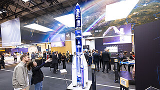 Modell einer Rakete auf einer Messe, umringt von Besucher*innen.