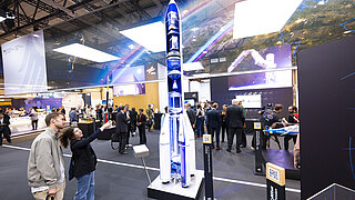 Model rocket on display at an exhibition, surrounded by visitors.