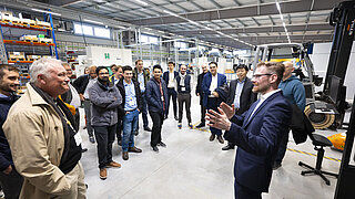 Ein Mann im Anzug spricht zu einer Gruppe von Zuhörenden in einer Fabrik.